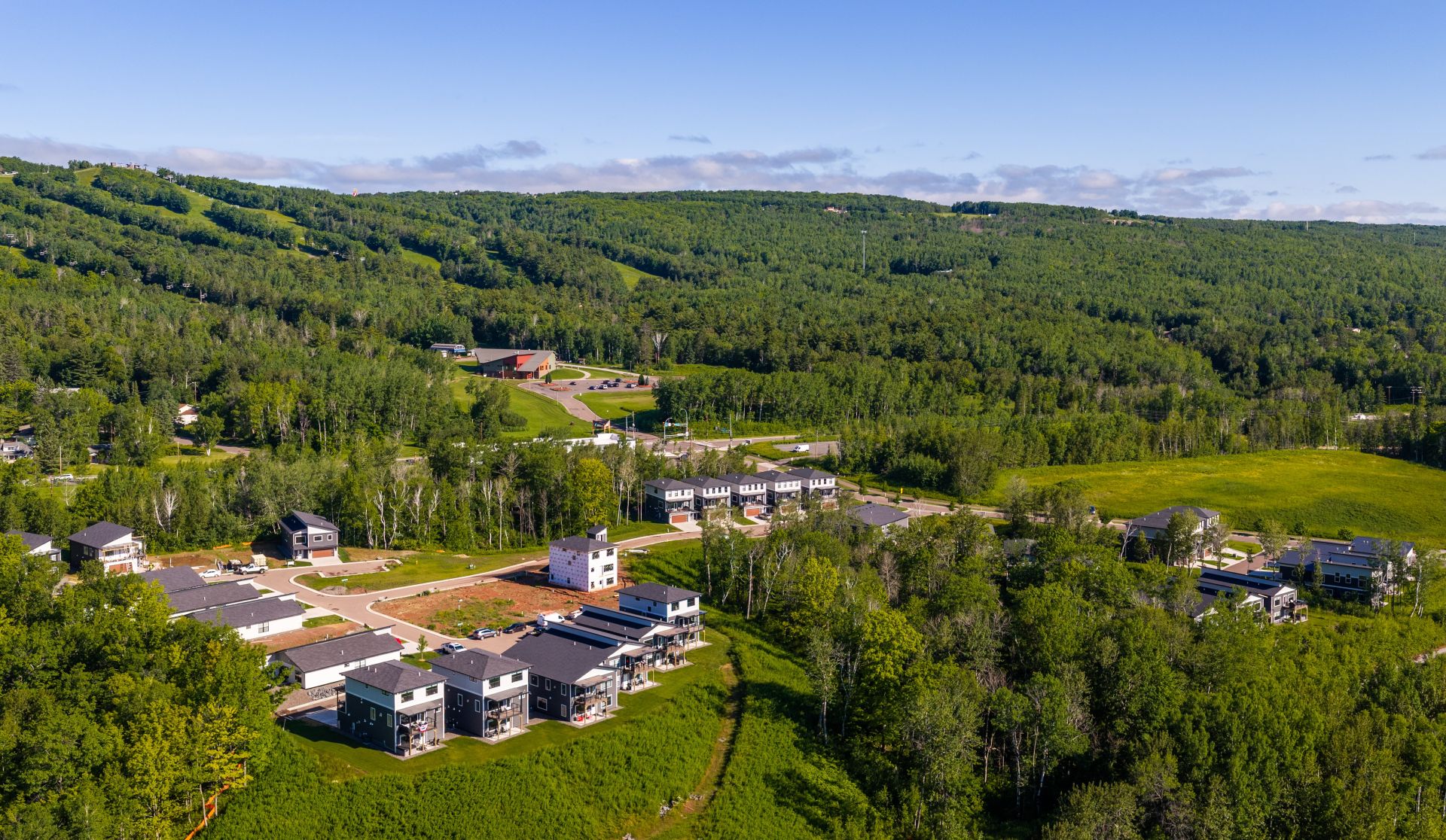 Aerial view of a suburban neighborhood bordered by dense woods and rolling green hills under a clear blue sky, with modern houses and roads in the foreground.