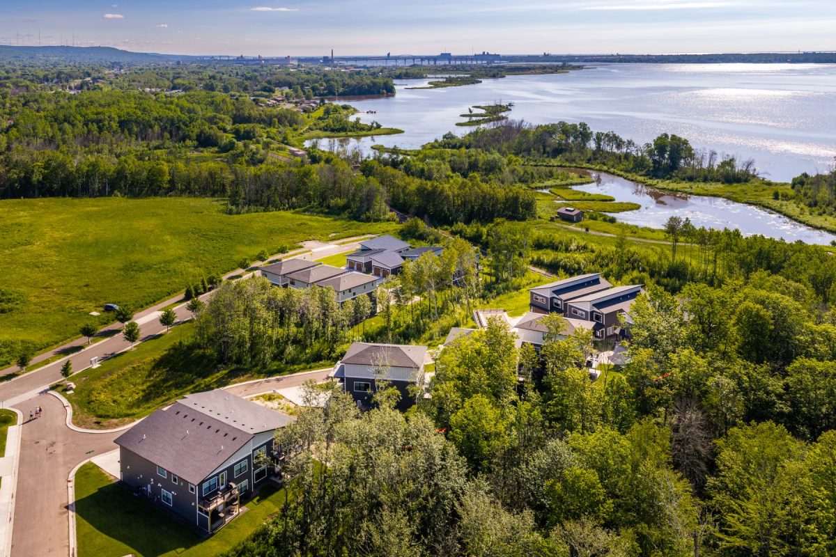 Aerial view of modern houses lining a curving street, with green lawns and dense trees beside a wide river.